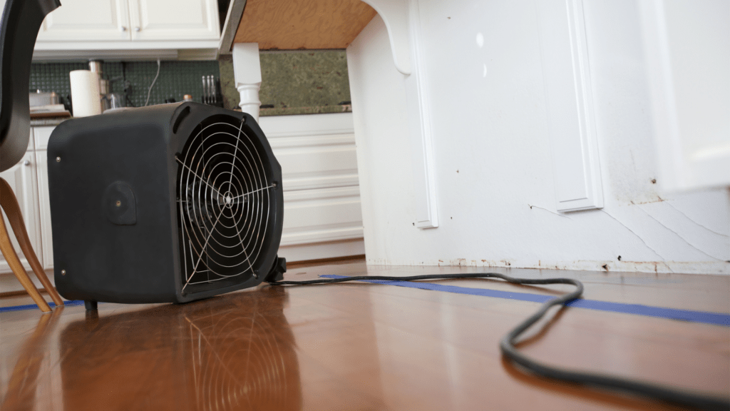 A black air mover fan on a hardwood floor aimed towards the base of white kitchen cabinets, suggesting an effort to dry out the area. Water damage is visible on the lower part of the cabinets, with staining and cracking paint.