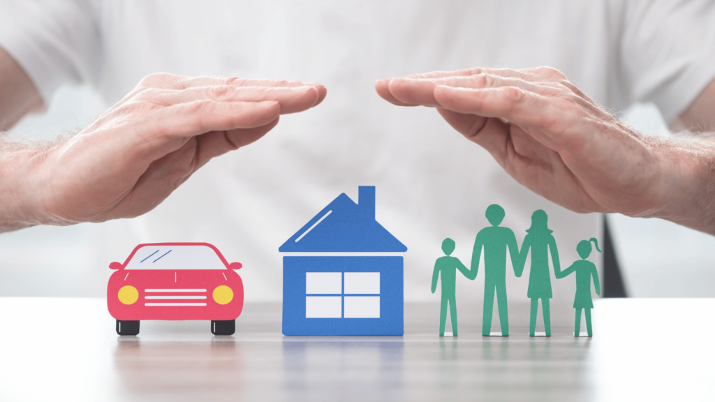 The image depicts two hands hovering protectively over representations of a red car, a blue house, and a green cut-out of a family with two adults and two children. It suggests themes of insurance, protection, and family safety.