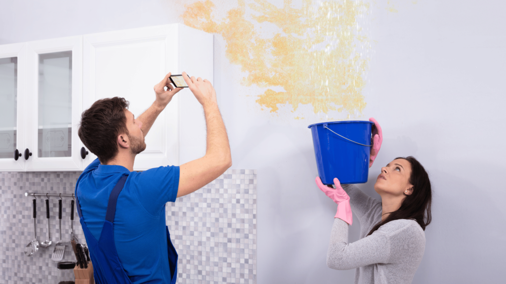 A woman is holding a bucket to collect water from a leaking ceiling, while a man is holding a phone to document the damage