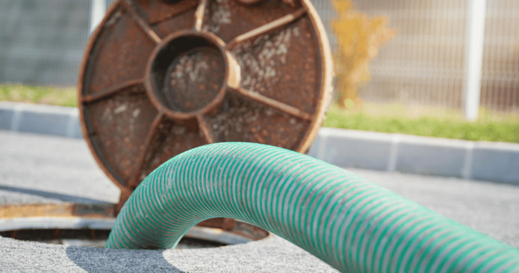 Close-up of a green hose being used to pump out a septic tank, with the rusty septic tank lid open in the background.