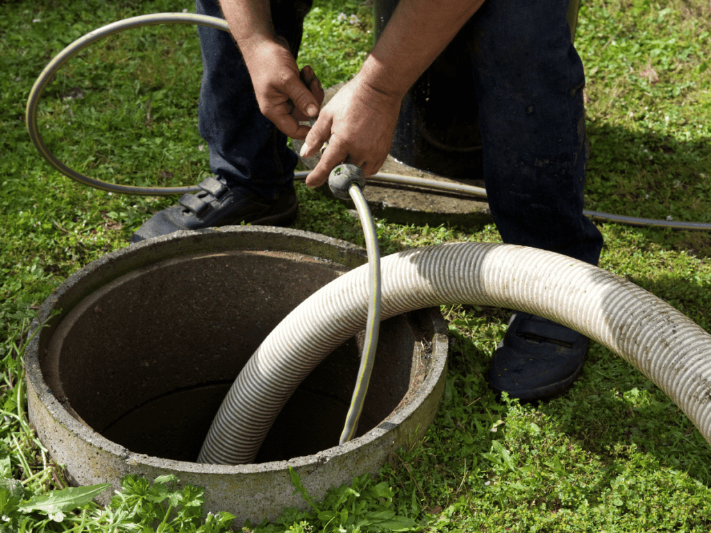 Worker cleaning a septic tank with a hose and vacuum equipment, focusing on the open septic tank lid and the worker's hands.